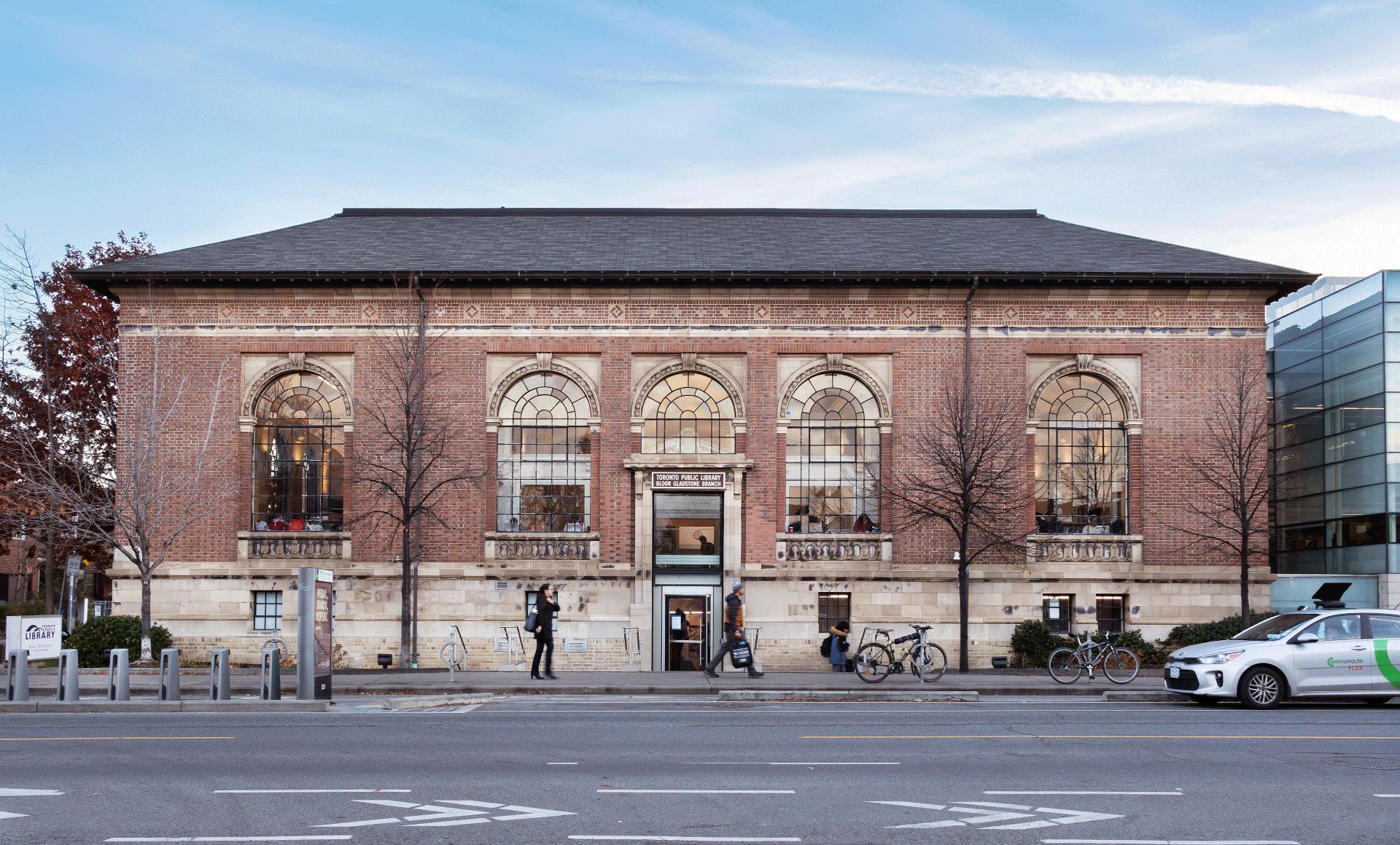 A red and cream brick building with a dark grey roof across a street. Three windows arch above the door. The sign above the door reads "Toronto Public Library Bloor Gladstone Branch".