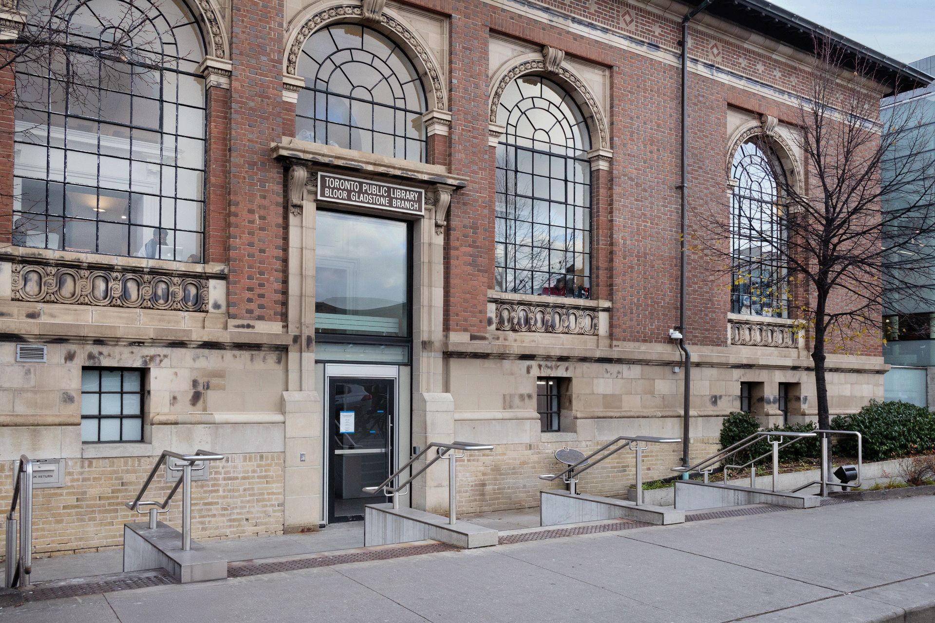 A close-up of a red and cream brick building with three arched windows. The sign above the door reads "Toronto Public Library Bloor Gladstone Branch".
