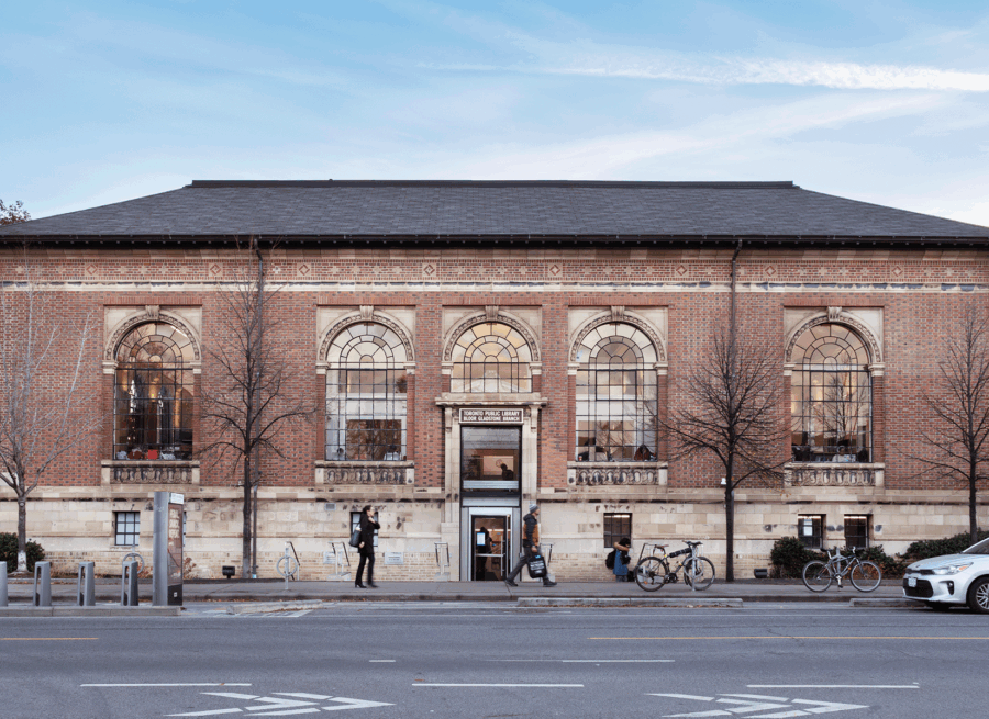 A red brick building with a dark grey roof across a street. Three windows arch above the door. The sign above the door reads "Toronto Public Library Bloor Gladstone Branch".