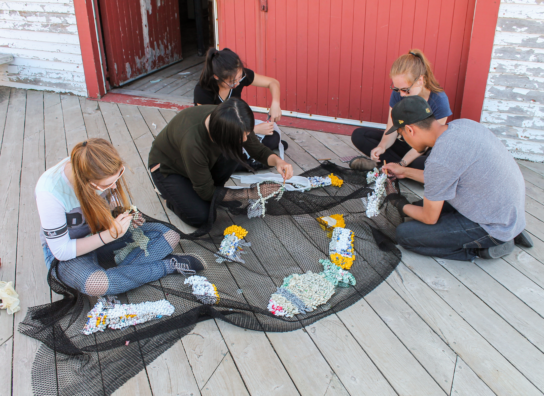 A group of people sitting on the ground weaving fish patterns into a fishnet.