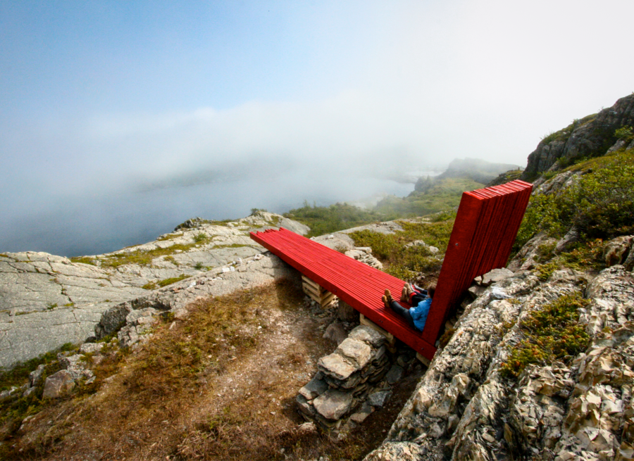 Large red wooden chair at hiking lookout, surrounding by greenery, rocks, and water below. 