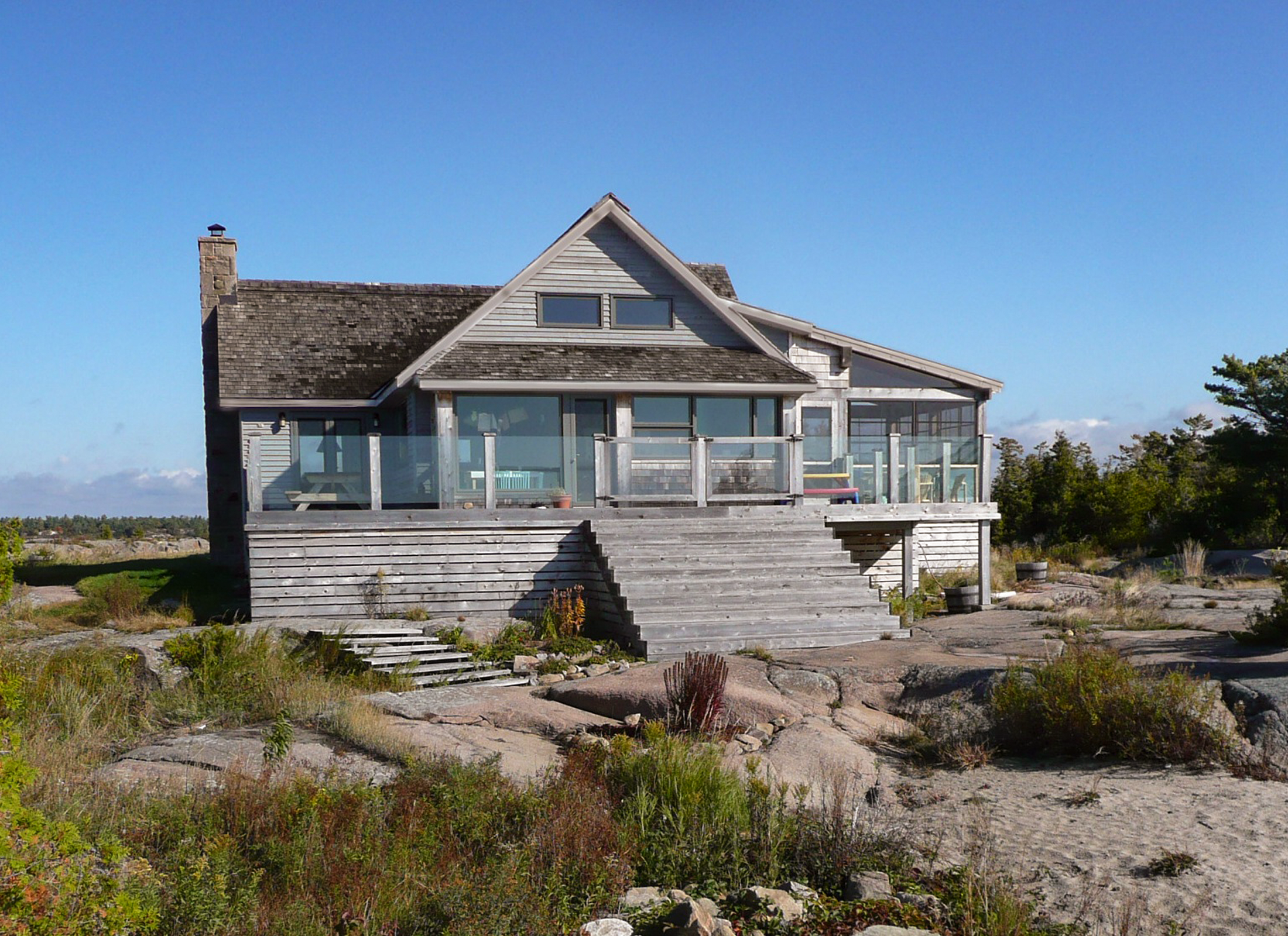 Exterior view of a cottage with grey wood siding, a large front stairway sitting on rocky terrain.