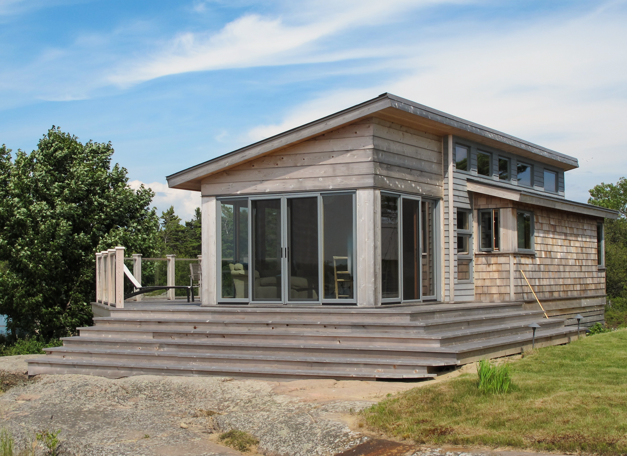 a bunkie with a slanted roof and glass sliding doors, with a patio on grass and rocky terrain.