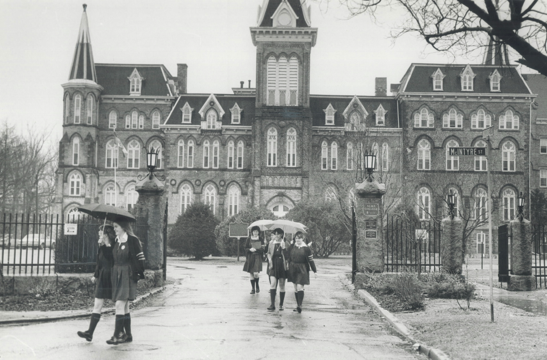 photo of teenage girls holding umbrellas walking by the entry gates of Alma College in 1983