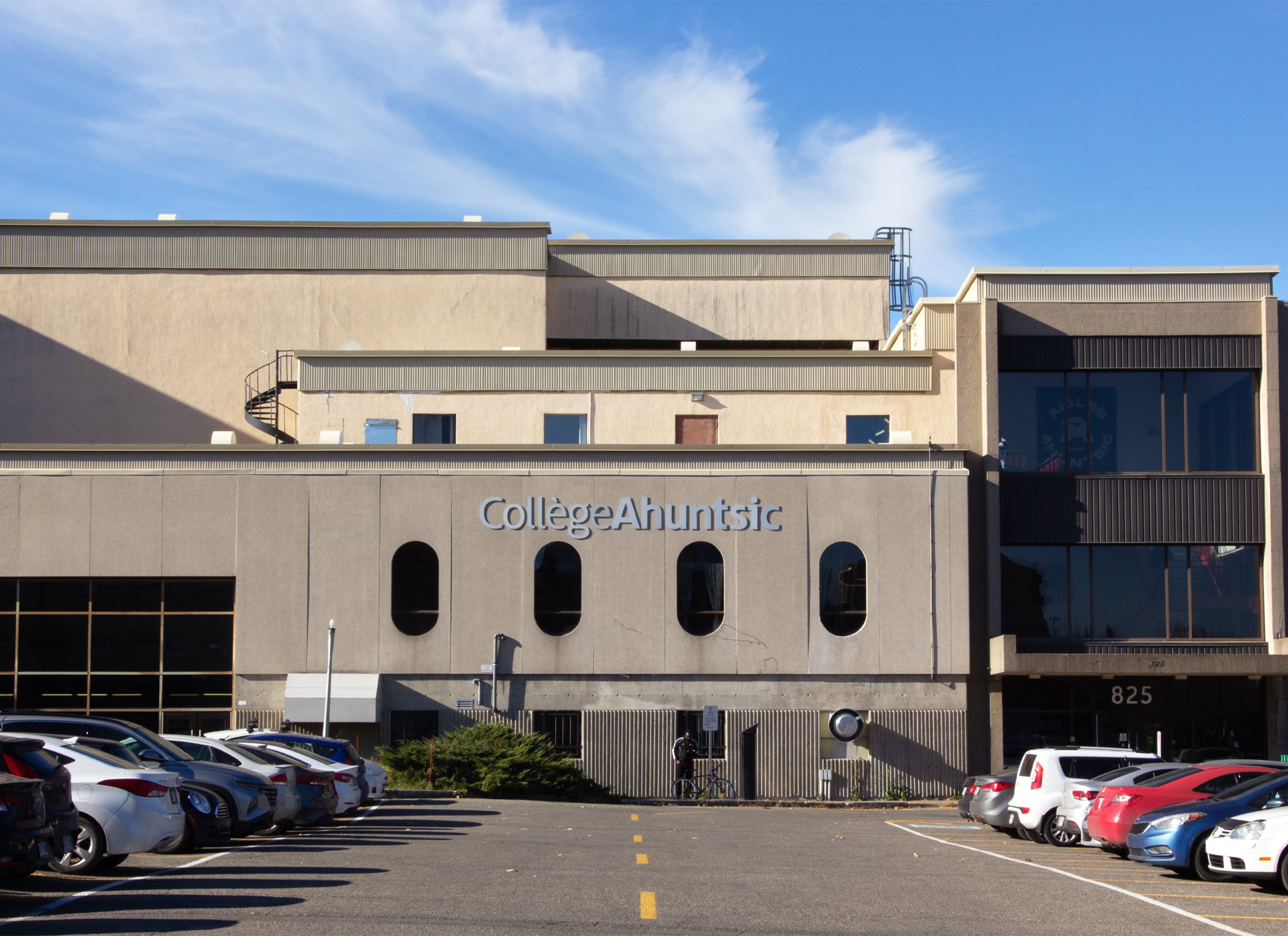 Exterior of Collège Ahuntsic with oval windows, and name on building. In foreground is a parking lot with cars.