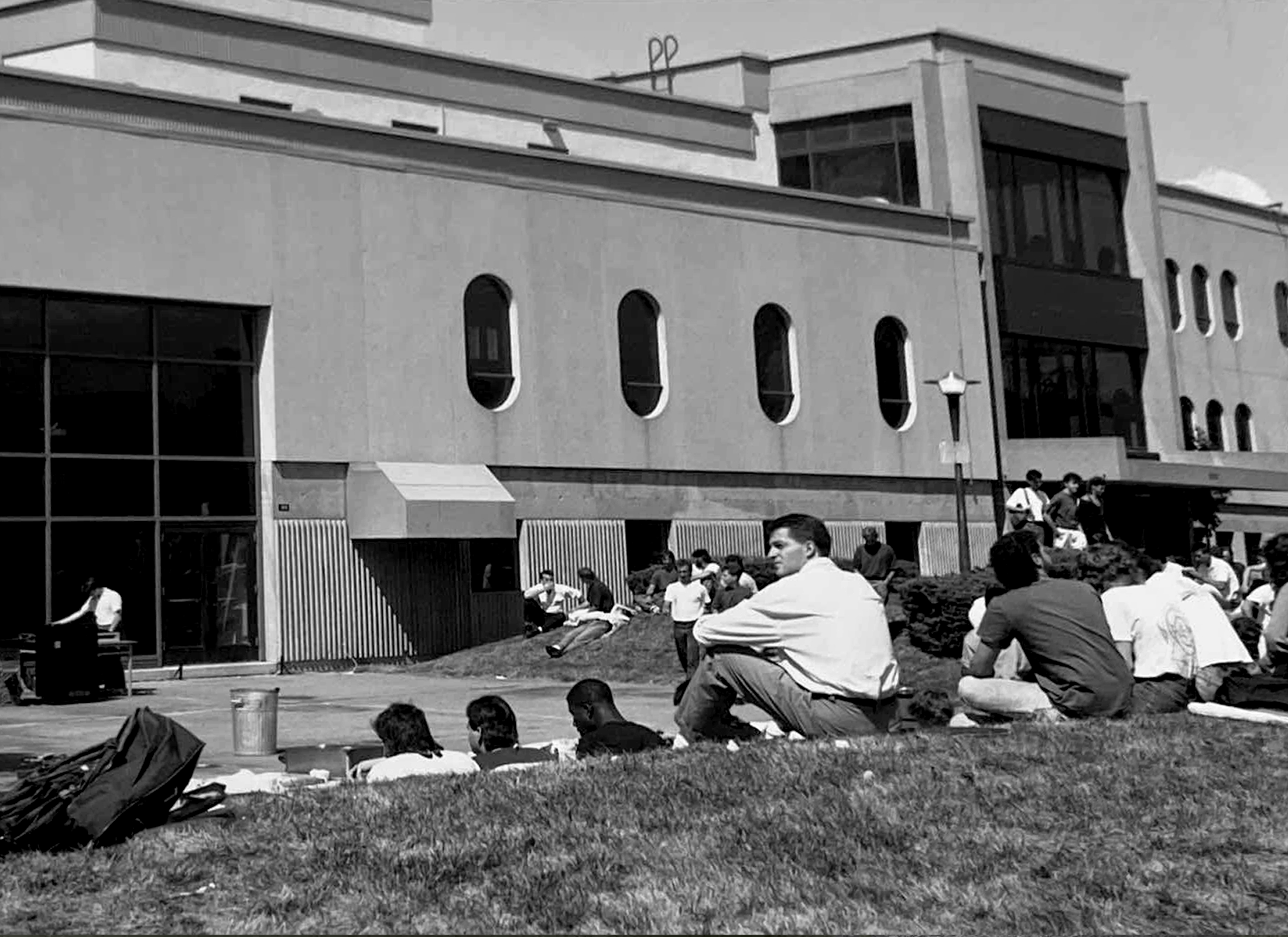 Group of people sitting on lawn in front of Collège Ahuntsic, a concrete building with oval windows.