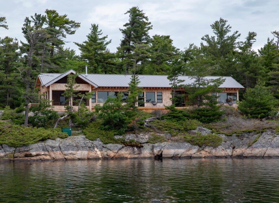 Cottage with wood siding and metal roof, with forest in background, and water in foreground.