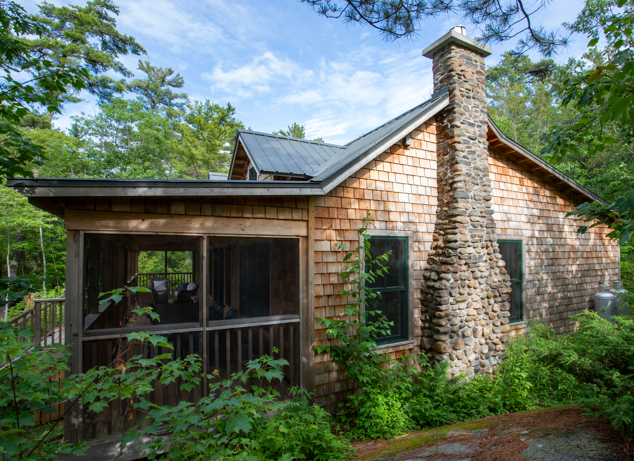 Exterior side view of Ballentine Pinewood cottage with cedar siding, black tin roof, large patio, dormer windows, and rock fireplace, settled into a lush green forest.