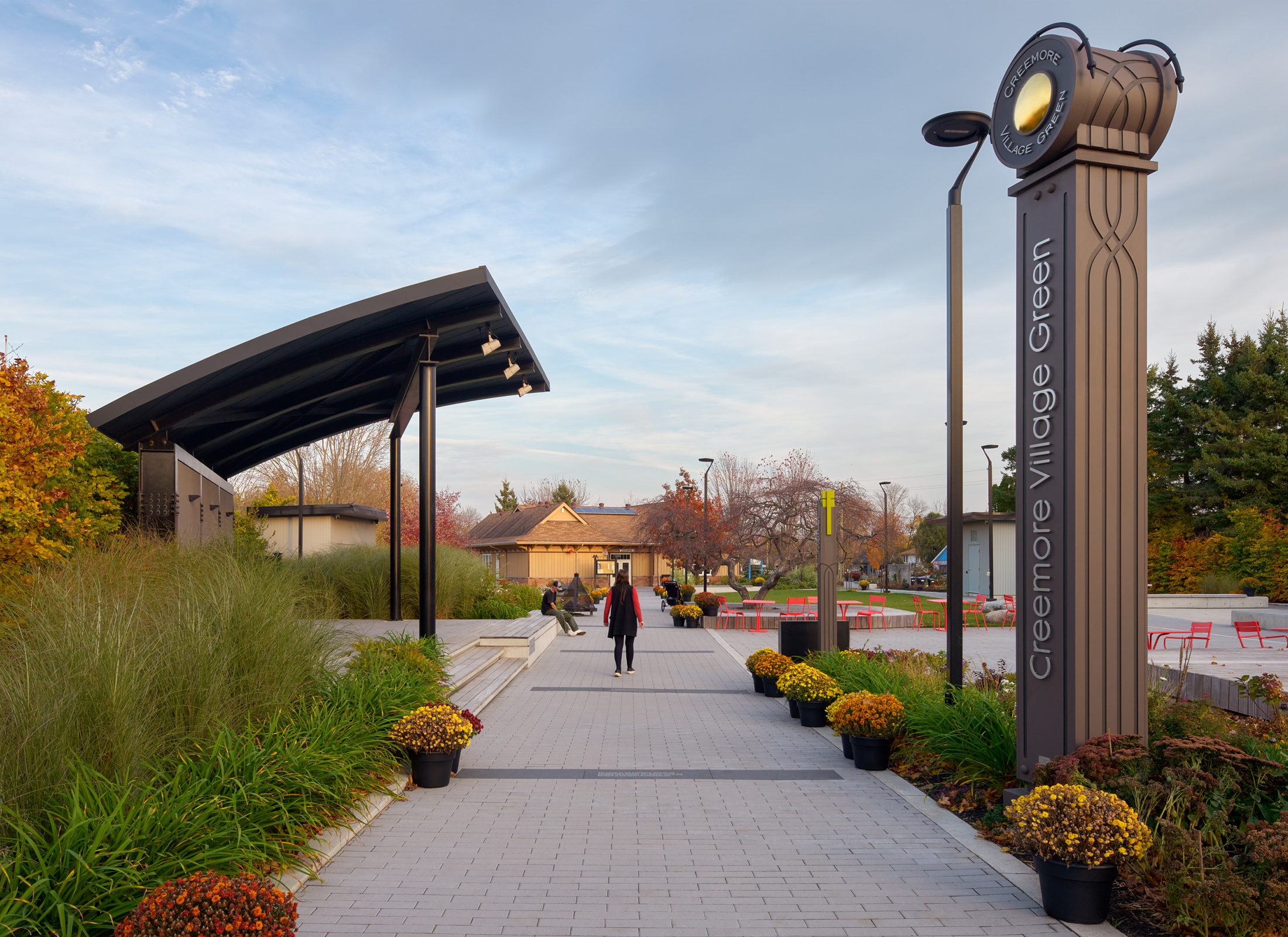 Pavilion with rounded roof surrounding by lush landscape, and red seating, and a tall sign that says "Creemore Village Green".