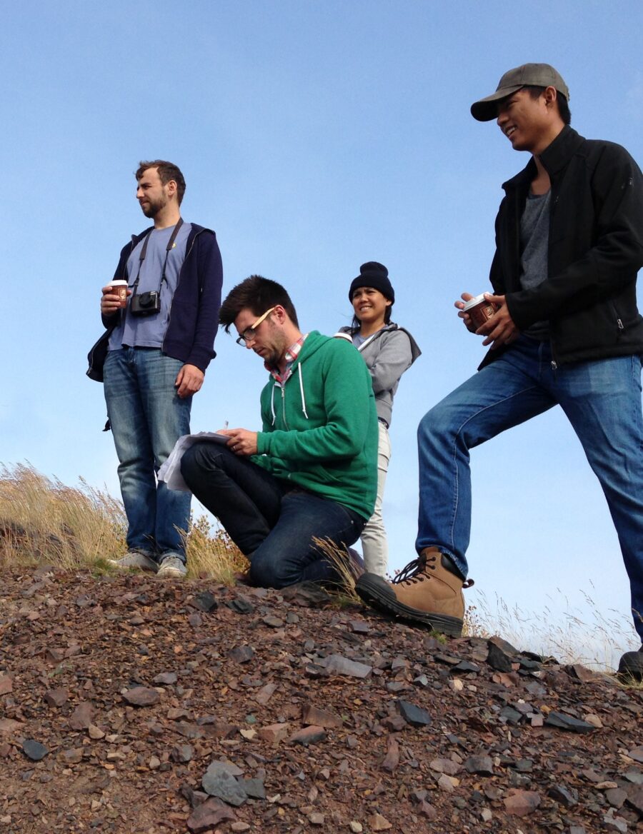 Philip Evans kneeling down writing on paper surrounded by three people on top of a hill, Bell Island.