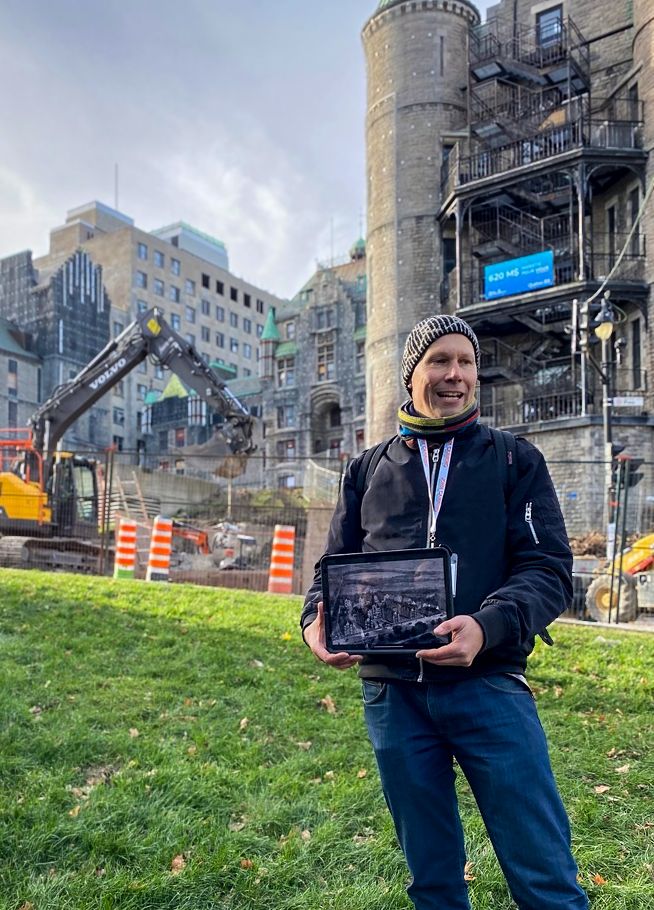 Jan Kubanek standing in font of the former he former Royal Victoria Hospital holding a tablet with an historical photo of the buildong behind him.