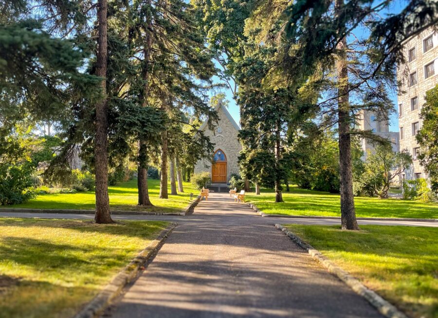 Gravel path leading to the Gardens, lush grass landscape, and heritage stone walls at the “Cité-des-Hospitalières" heritage building, lined with trees and benches.